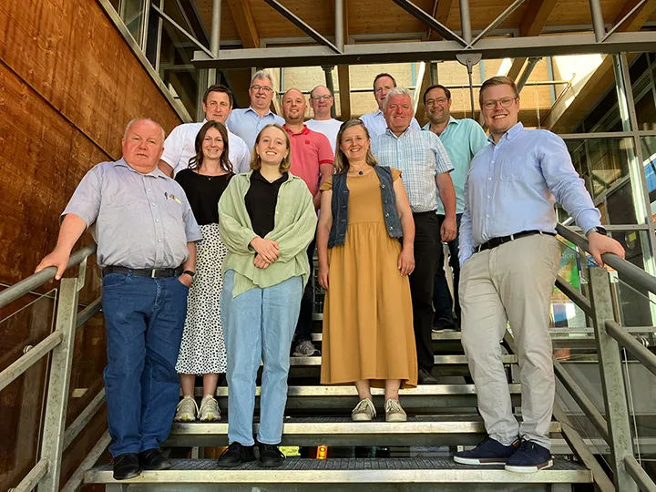 Gruppenfoto von 14 Personen auf einer Treppe in einem modernen Gebäude mit Glasfassade und Holzverkleidung – Teilnehmerinnen und Teilnehmer stehen in zwei Reihen und blicken in die Kamera
