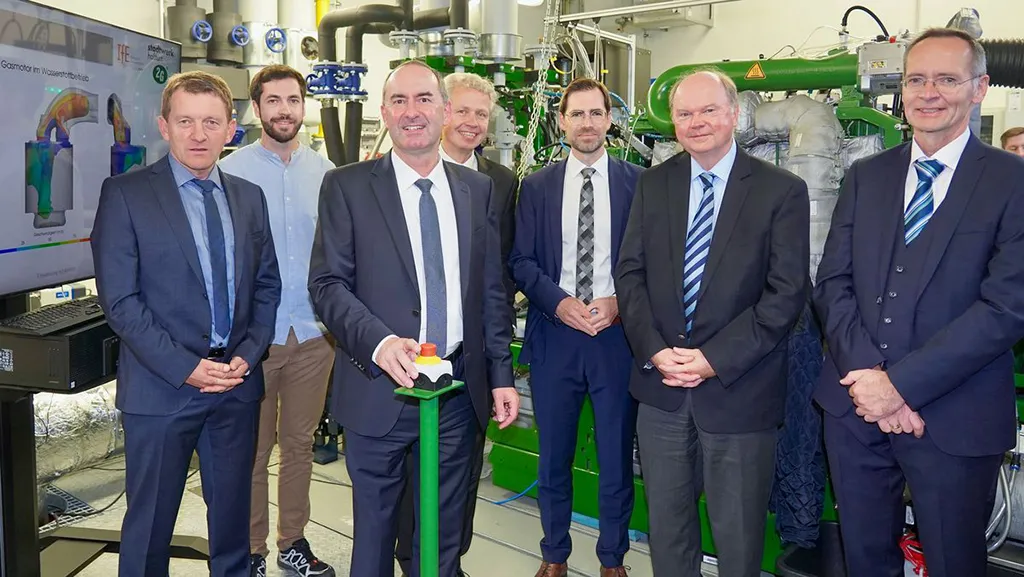 Gruppenfoto von acht Personen in einem technischen Raum mit grünem Blockheizkraftwerk im Hintergrund – eine Person betätigt symbolisch einen Startknopf, auf einem Monitor ist ein Gasmotor-Modell zu sehen