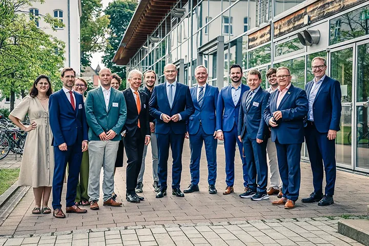 Gruppenfoto von 14 Personen in Businesskleidung vor einem modernen Gebäude mit Glasfassade – aufgenommen im Außenbereich auf einem gepflasterten Weg