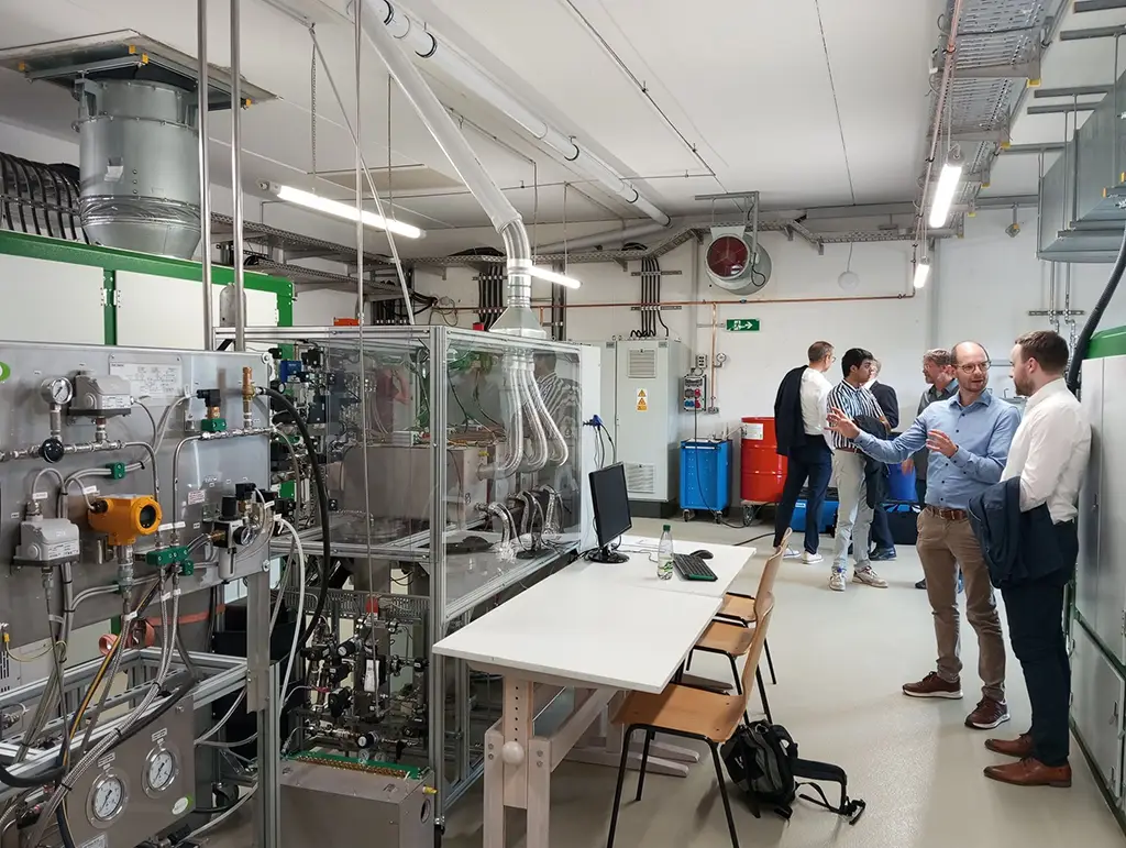 Innenaufnahme eines Technikraums mit komplexer Anlagentechnik und mehreren Personen im Gespräch – moderne Laborumgebung mit Monitor, Rohrleitungen und Steuerungssystemen